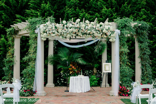 outdoor ceremony mandap,white floral arch,greenery wedding decor,elegant ceremony setup,garden wedding venue,mandap design,wedding aisle decor,romantic outdoor setting,floral installation,ceremony backdrop