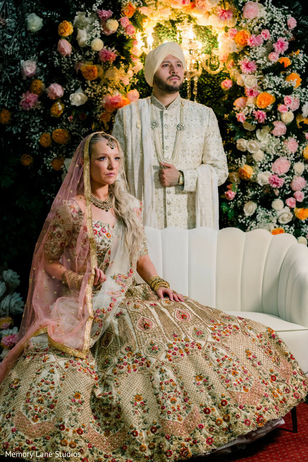 bridal lehenga embroidery,groom ivory sherwani,floral backdrop portrait,regal couple pose,luxury wedding decor,bride seated portrait,groom standing portrait,multicultural wedding style,elegant couple styling,reception portrait