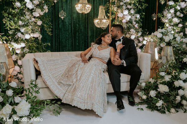 reception couple portrait,floral stage decor,bride ivory lehenga,groom black tuxedo,romantic sofa moment,elegant wedding stage,luxury wedding decor,couple candid interaction,indoor reception styling,greenery backdrop