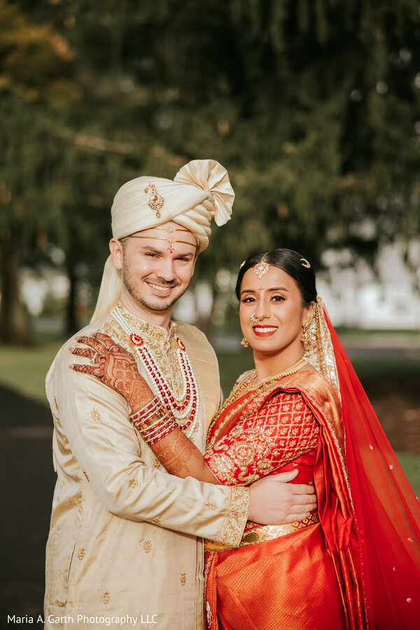 indian wedding couple portrait,bride orange saree,groom cream sherwani,outdoor wedding photo,couple embrace pose,traditional indian attire,bridal jewelry details,groom turban style,romantic couple shot,cultural wedding portrait