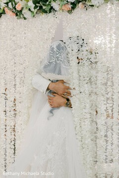 Couple Embracing Against Floral Backdrop in White Wedding Attire