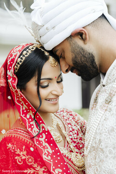 This close-up portrait features a couple in traditional wedding attire, with the bride wearing a red lehenga and the groom in a white sherwani and turban. They are shown in a tender moment, heads touching, showcasing intricate bridal jewelry and a feathered turban.