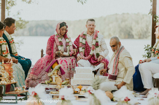 outdoor ceremony,pink lehenga,floral garlands,wedding ceremony decor,white groom attire,lake view,traditional ceremony setup,wedding rituals,bridal fashion,groom fashion