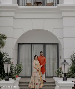 Couple in Floral Lehenga and Orange Kurta Against White Archway