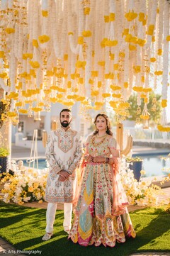 A couple stands beneath a canopy of white and yellow floral strings. The bride wears a light blue and green lehenga with intricate colorful patterns, and the groom wears a white sherwani with embroidered details.
