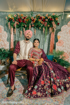 Couple in Plum Lehenga and Plum Kurta Seated Before Floral Backdrop