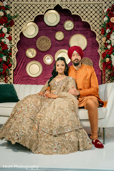 couple portrait,gold lehenga,gold kurta,red turban,floral decor,stage backdrop,wedding fashion,maroon backdrop,decorative wall hangings