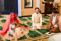 Bride in Gold Attire and Red Dupatta with Groom in Cream Sherwani Kneeling