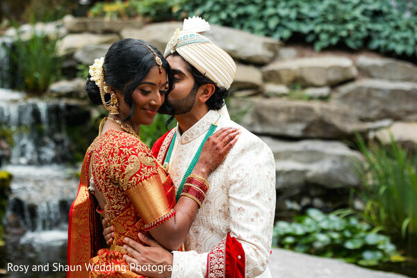 couple portrait,wedding attire,red sari,cream sherwani,outdoor wedding,water feature,romantic pose,wedding fashion,groom kissing bride