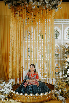 Woman in Red and Blue Lehenga Seated in Golden Lotus Structure Amidst Yellow Floral Decor